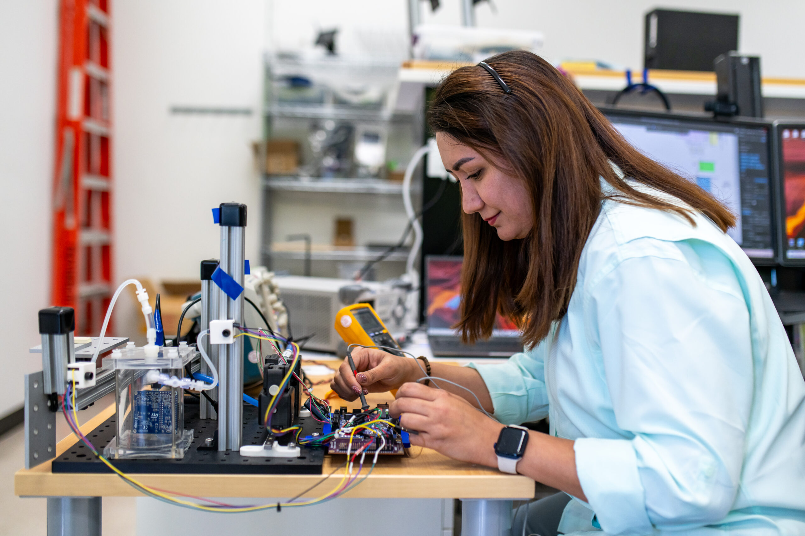 A woman wearing a light blue shirt and a smartwatch is seated at a workbench, focused on working with electronic components and circuit boards. She is using probes to test a circuit connected with colorful wires, alongside lab equipment and instrumentation. In the background, computer monitors display data and diagrams, and shelves hold various equipment and tools in a well-organized technical workspace.
