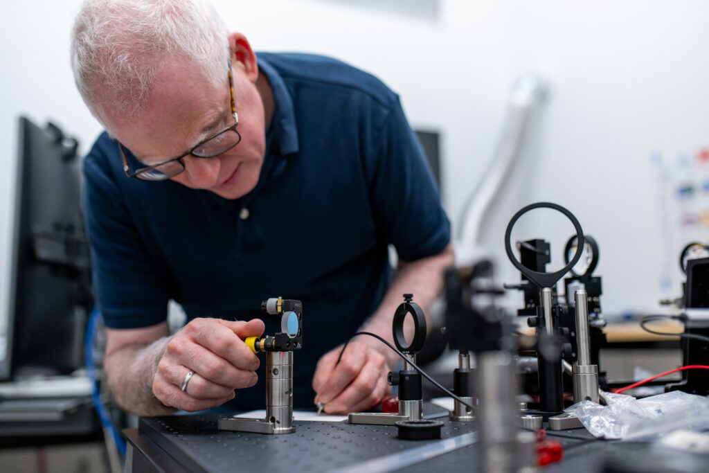 An older man with white hair and glasses, wearing a navy blue shirt, is carefully adjusting a precision optical instrument on a lab bench. He is using a small tool to fine-tune the setup, which includes mirrors, lenses, and metal mounts arranged on a perforated work surface. The background shows computer equipment and lab tools, indicating a highly technical and focused laboratory environment.