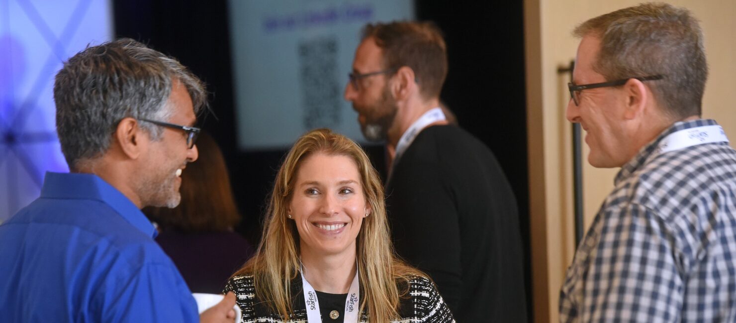 A smiling woman holding a coffee mug chats with two male attendees during a networking break. All wear StarFish Medical lanyards and conference badges. The setting is informal and social, with other attendees conversing in the background.