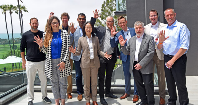 A group of speakers at Medical Device Playbook Newport Beach 2025 pose outdoors on a terrace with palm trees and a golf course in the background. Most are waving with one hand raised. They are dressed in business casual and formal attire, suggesting a networking or conference setting.