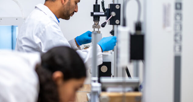 Two scientists wearing lab coats and blue gloves working with precision instruments in a laboratory setting, focusing on mechanical or materials testing equipment.