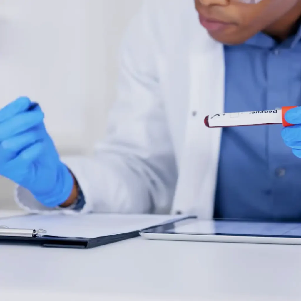 ELLA AI FDA regulatory research tool ELLA AI FDA regulatory research tool A scientist wearing blue gloves and a lab coat is holding a blood sample tube labeled “Dengue” while taking notes on a clipboard. The background shows a sterile lab environment.