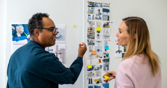 Two colleagues collaborating in front of a whiteboard covered with printed images and sticky notes, discussing design concepts or project ideas.