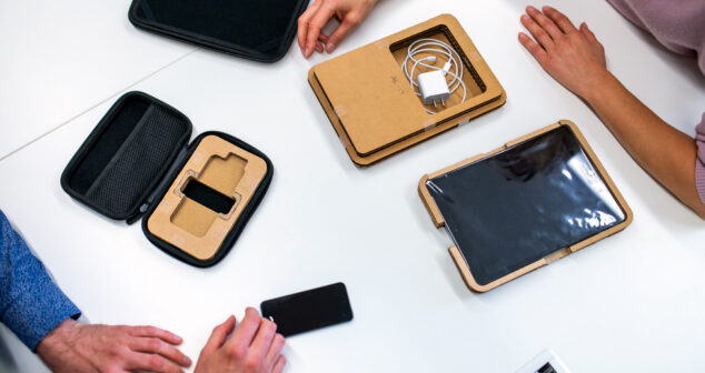 Top-down view of three people at a white table examining electronic device packaging, including tablets, chargers, and protective cases.