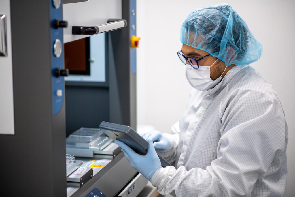 Startups & Founders A person working in a cleanroom environment, wearing a white lab coat, blue hairnet, protective face mask, safety glasses, and blue gloves. They are operating or inspecting equipment, holding a tablet or control device, with laboratory machinery and trays visible in the background. The setting appears to be a sterile lab or manufacturing facility.
