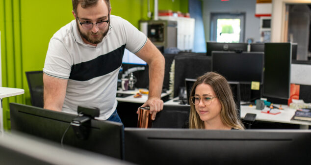A man standing next to a seated woman, both looking at dual computer monitors in a modern office with green walls and multiple desks in the background.