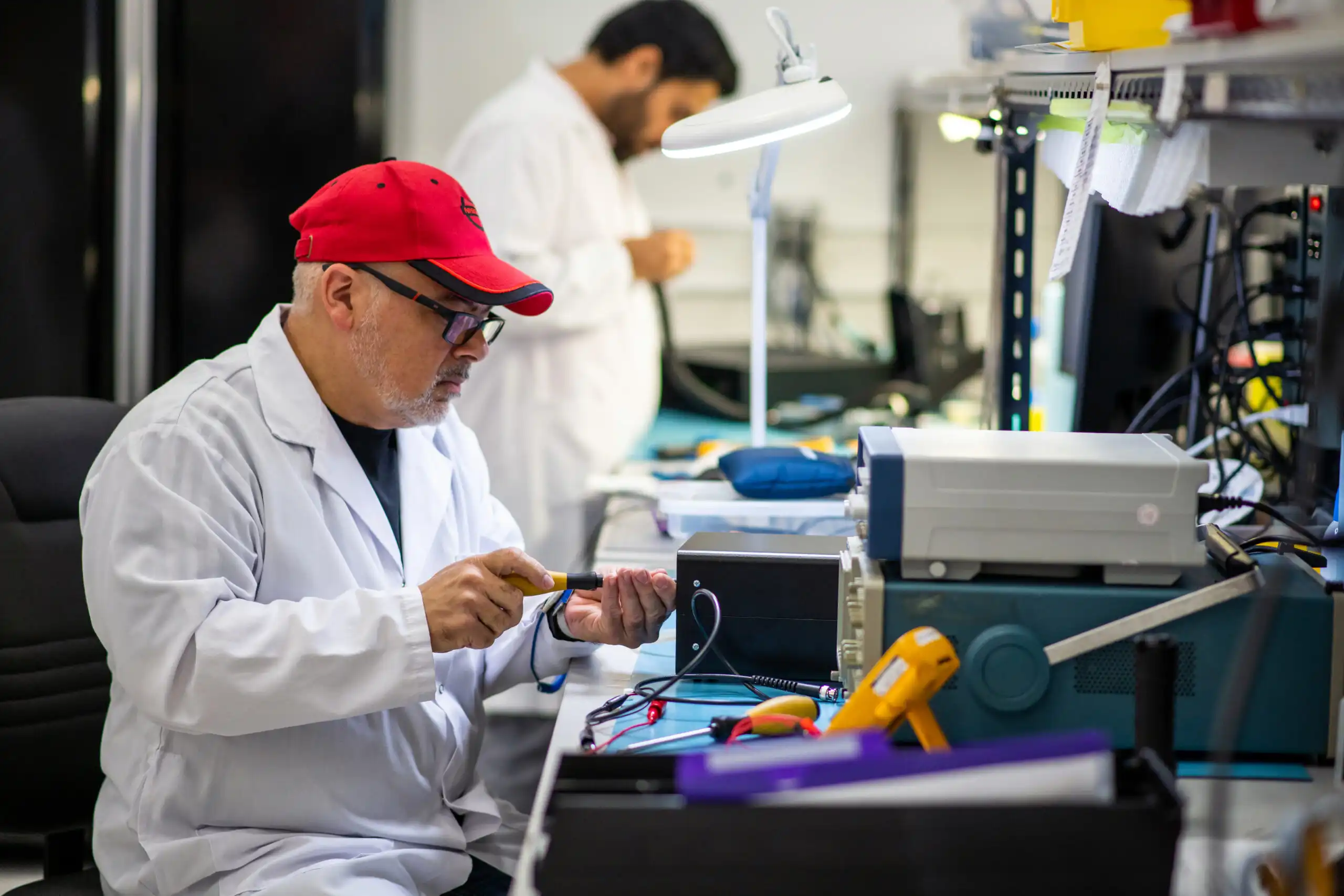 new product introduction Startups & Founders new product introduction Two technicians working in a laboratory or electronics workshop. In the foreground, a man wearing a white lab coat, red baseball cap, and glasses is seated at a workbench, using a tool to work on an electronic device. The bench is cluttered with various instruments, tools, and equipment. In the background, another technician in a white lab coat is standing and working at a separate station under a bright task light. The environment is organized and technical, indicating detailed assembly or testing work.
