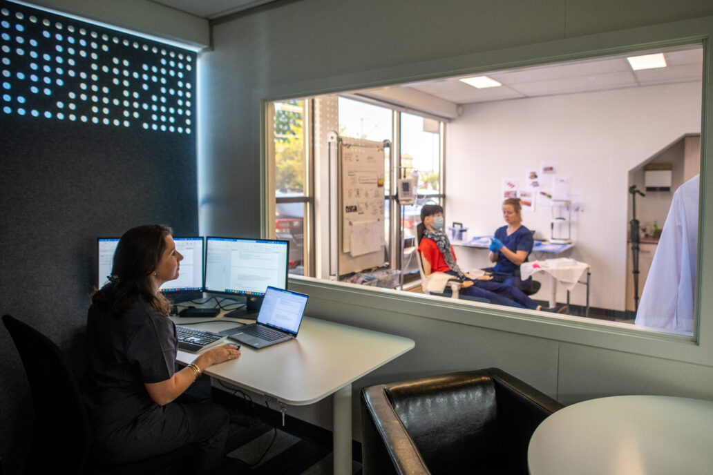 The image shows a professional workspace divided by a glass window. In the foreground, a person is seated at a desk with multiple computer monitors and a laptop, seemingly observing or recording data. Through the glass window, two individuals are visible in a clinical or lab-like setting. One is seated in a chair, wearing a mask and scarf, while the other, dressed in scrubs and gloves, appears to be conducting a procedure or simulation. The environment suggests a usability testing scenario.
