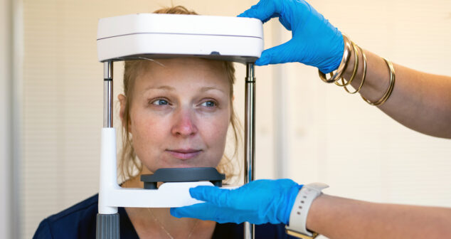 A woman sits still while a medical professional wearing blue gloves adjusts a diagnostic imaging or eye examination device positioned around her face.