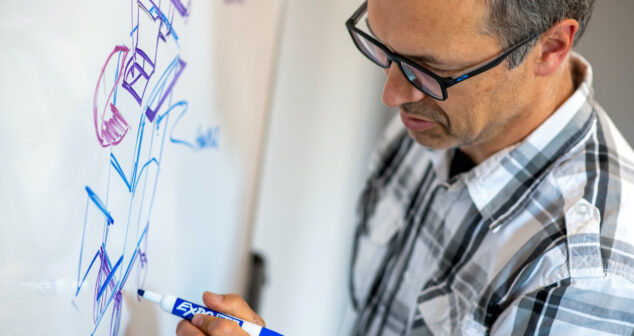 A man wearing glasses and a plaid shirt draws on a whiteboard with a blue Expo marker. The whiteboard contains various colorful sketches and diagrams, likely representing technical or design concepts. The man is focused on his drawing, appearing deep in thought.
