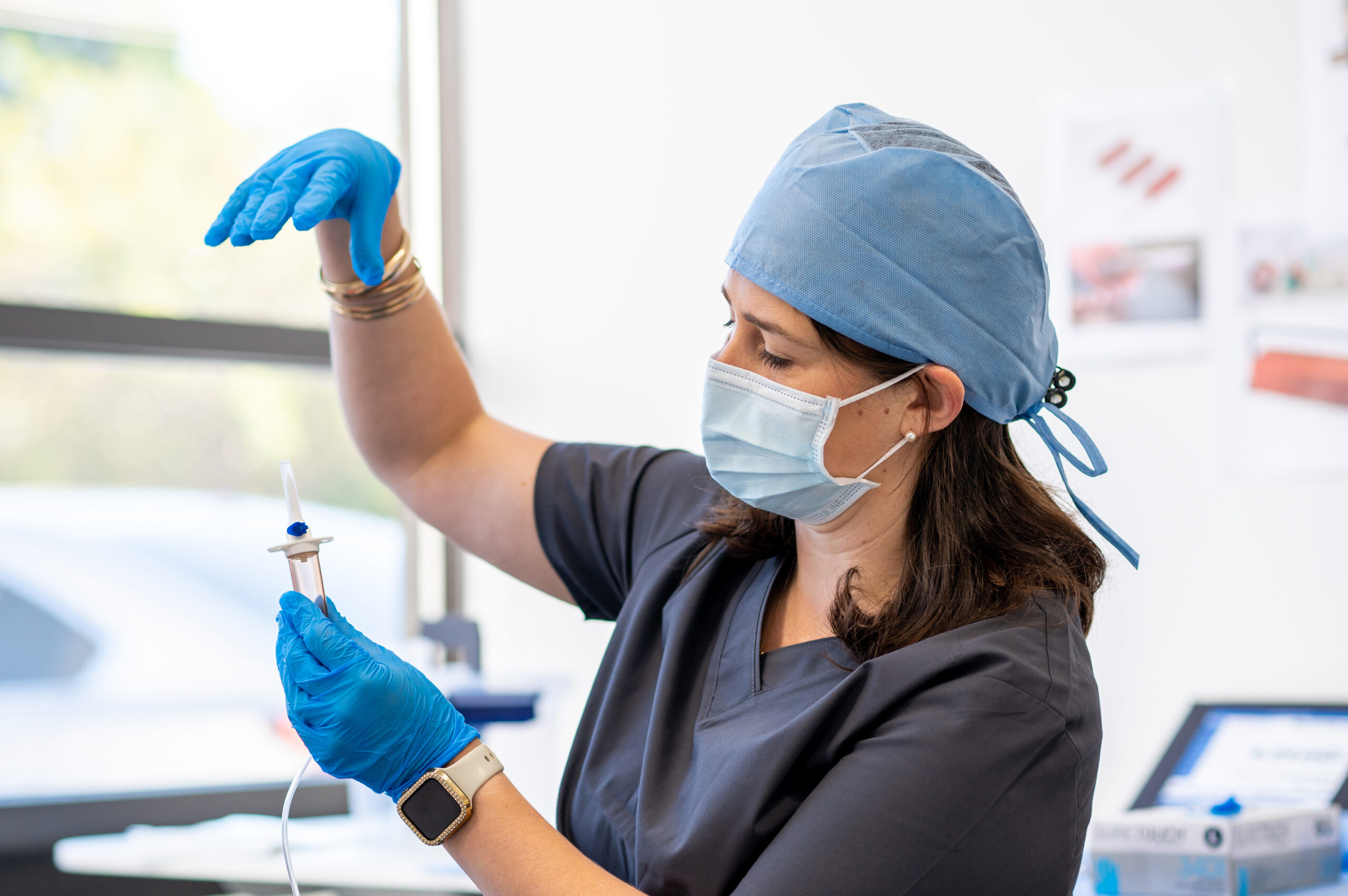 Startups & Founders A healthcare professional wearing scrubs, a blue surgical cap, a face mask, and blue gloves holds a medical device component, inspecting it carefully with both hands. She appears focused and precise, standing in a bright, clinical environment with medical supplies and images on the wall in the background.
