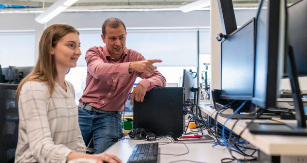 Engineer guiding a colleague through software on dual monitors in a device lab
