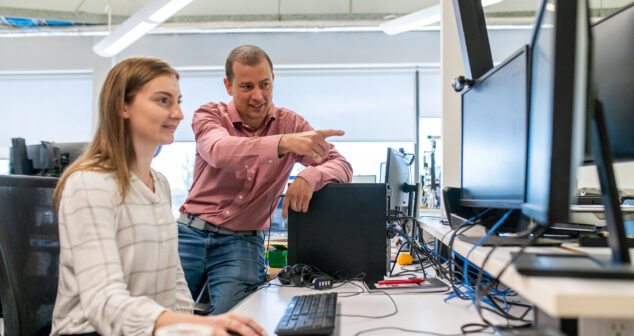 A man standing and pointing at a computer screen while a seated woman uses a mouse and keyboard, both focused on a task in a modern office or lab workspace.