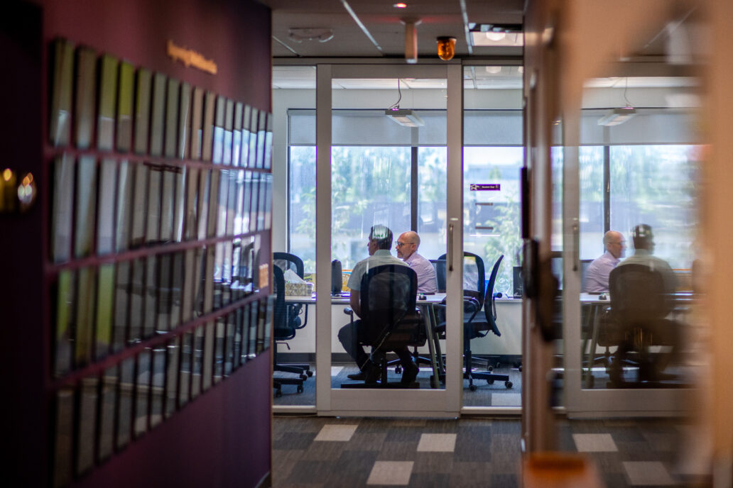 careers at StarFish Medical A hallway view into a modern office meeting room with glass walls. Inside, three people are seated around a conference table, engaged in conversation. The room has large windows with daylight streaming in, and office chairs around the table. On the left side of the hallway, there is a purple wall with rows of small plaques or awards mounted on it.