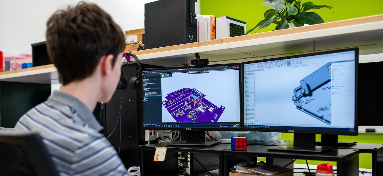 A person wearing a striped shirt sits at a desk with dual monitors, working on a CAD design of a circuit board and a 3D model. The workspace includes a headset, a Rubik's Cube, a plant, and various equipment.