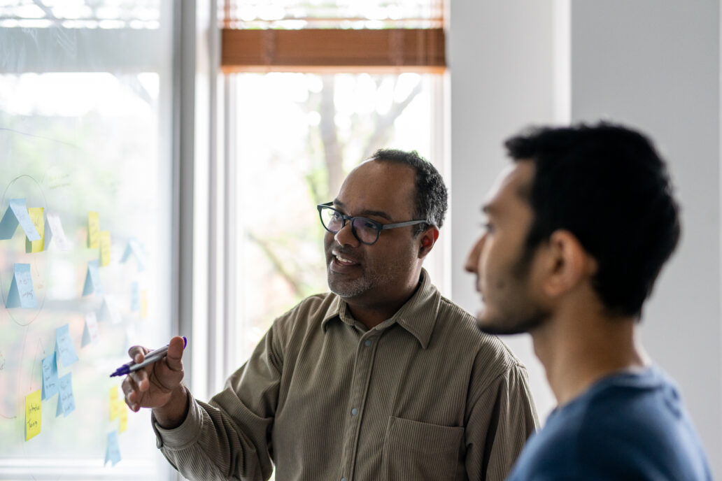Two men standing in front of a window and whiteboard covered with colorful sticky notes. One man, wearing glasses and a brown shirt, holds a purple marker and gestures toward the notes while explaining something. The other man, in a blue shirt, listens attentively. Natural light streams in through the window, creating a bright and collaborative atmosphere.