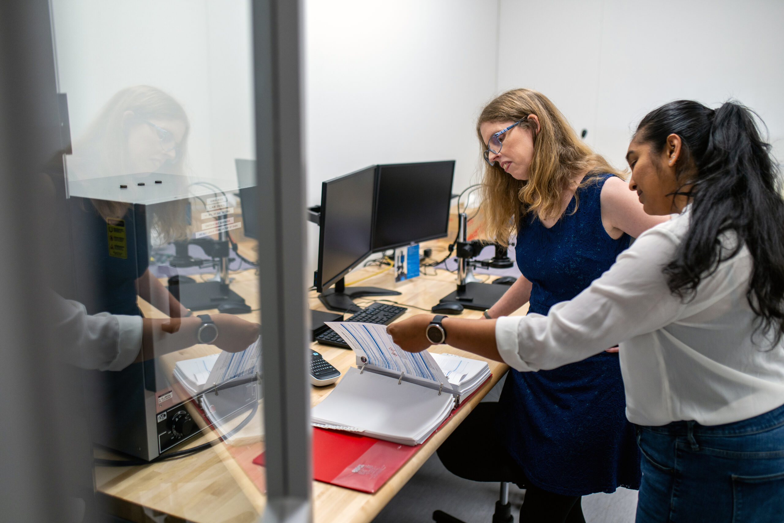 Startups & Founders Two women working together in an office or lab setting. One woman, wearing a blue dress and safety glasses, sits at a desk with dual computer monitors, while the other woman, in a white blouse and jeans, stands beside her, pointing to documents in an open binder on the desk. The scene is viewed through a glass partition, with reflections partially visible, and the workspace includes technical equipment and office supplies.