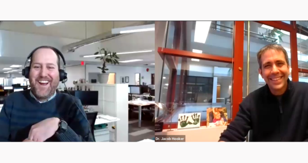 A screenshot from a virtual webinar or video call hosted by StarFish Medical. On the left, a man labeled 'Nick Allan' is wearing a headset and laughing, sitting in an open office space with multiple computer monitors and employees in the background. On the right, a man labeled 'Dr. Jacob Hooker' is smiling while sitting in an office with large windows, framed pictures, and artwork behind him.