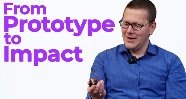 A man in a blue shirt and glasses, wearing a lapel microphone, smiles while holding a small circular device with wires. Large bold purple text on the left side of the image reads "From Prototype to Impact" against a white background.
